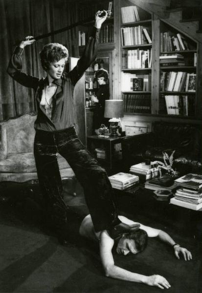 Scena del film "La donna invisibile" - Regia Paolo Spinola, 1969 - Figura intera di Carla Gravina con un piede sulla testa di Silvano Tranquilli. L'attrice, agita in aria la cintura dei pantaloni di lui che è sdraiato prono a torso nudo.