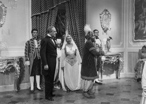 Scena del film "La donna perduta" - Regia Domenico Gambino, 1940 - Alberto Capozzi, in piedi accanto a Carlo Campanini in abiti orientali. Intorno a loro, attori e attrici non identificati.