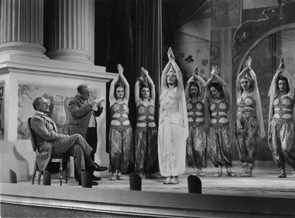 Scena del film "La donna perduta" - Regia Domenico Gambino, 1940 - Su un palcoscenico Elli Parvo tra un gruppo di ballerine, tutte vestite in abiti orientali. Seduto a sinistra, Alberto Capozzi le guarda. Accanto a lui un attore non identificato.