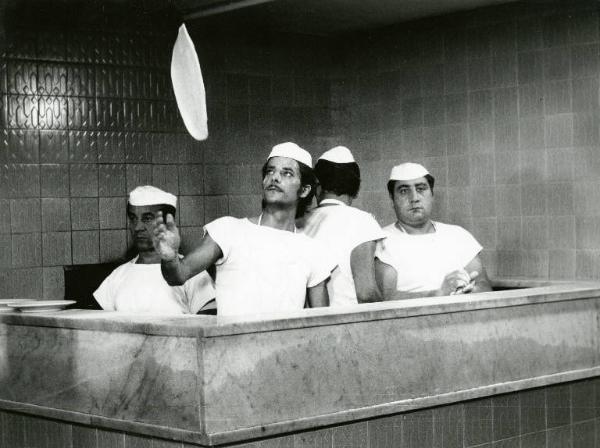 Scena del film "Dramma della gelosia (tutti i particolari in cronaca)" - Regia Ettore Scola, 1970 - Giancarlo Giannini prepara la pasta per pizza dietro un banco di pizzeria. Intorno a lui, attori non identificati nei panni di altri pizzaioli.