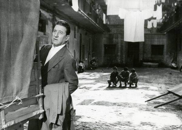 Scena del film "I due compari" - Regia Carlo Borghesio, 1955 - Carlo Ninchi, in piedi a lato di un furgone, guarda dritto davanti a sé. Sullo sfondo tre bambini accovacciati giocano.