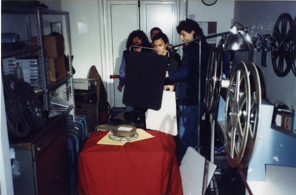 Sul set del film "Due dollari al chilo" - Regia Paolo Lipari, 2000 - Paolo Lipari e Matteo Pavesi, in secondo piano, insieme a due operatori non identificati della troupe, si preparano a riprendere una bobina di pellicola appoggiata a un tavolo.