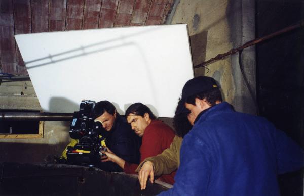 Sul set del film "Due dollari al chilo" - Regia Paolo Lipari, 2000 - Due membri della troupe manovrano la macchina da presa mentre, al centro della scena, il regista Paolo Lipari mostra a un operaio cosa deve fare.