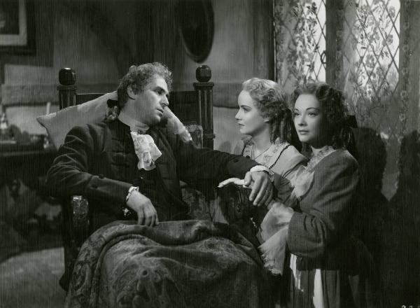 Scena del film "Le due orfanelle" - Regia Carmine Gallone, 1942 - Otello Toso, seduto su una poltrona a sinistra, si scambia un intenso sguardo con Alida Valli mentre tiene le mani di María Denis che guarda davanti a sé.