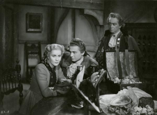 Scena del film "Le due orfanelle" - Regia Carmine Gallone, 1942 - Alida Valli, seduta con le braccia conserte a un tavolo mentre guarda dritto davanti a sé. Roberto Villa, a destra, la osserva. Alle loro spalle, in piedi, un attore non identificato.