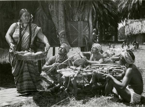 Scena del film "Le due tigri" - Regia Giorgio Candido Simonelli, 1941- Sulla sinistra, figura intera di Agnese Dubbini con una cesta in mano. L'attrice viene osservata da tre attori non identificati seduti a terra che lavorano.