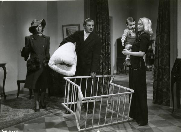 Scena del film "È caduta una donna" - Regia Alfredo Guarini, 1941 - A destra, Isa Miranda con un bimbo in braccio. Al centro, Claudio Gora tiene una culla e una coperta tra le mani. Dietro, Carla Martinelli sorride, con un cappello da uomo in mano.