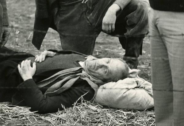 Scena del film "E cominciò il viaggio nella vertigine" - Regia Toni De Gregorio,1974 - Gastone Moschin, a terra supino con il capo poggiato su un sacco e le mani sul ventre, volge lo sguardo a destra. Chinato su di lui, un attore non identificato.