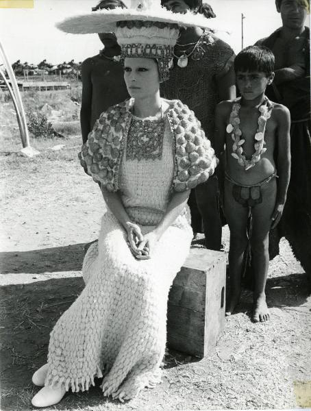 Scena del film "Edipo Re" - Regia Pier Paolo Pasolini, 1967 - Silvana Mangano, in abiti di scena, è seduta su una cassa di legno e rivolge lo sguardo verso sinistra. Dietro di lei, sono presenti quattro attori non identificati.