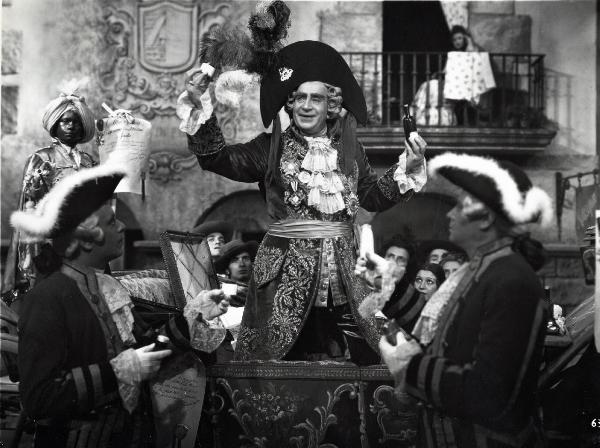 Scena del film "Elisir d'amore" - Regia Amleto Palermi, 1941 - Al centro, con un cappello decorato con piume, Armando Falconi mostra ad attori non identificati una bottiglietta di vetro che tiene nella mano sinistra e un altro oggetto.