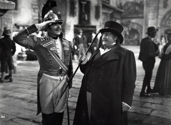 Scena del film "Elisir d'amore" - Regia Amleto Palermi, 1941 - A sinistra, un attore non identificato, sorride e fa il saluto militare a un altro attore non identificato, a destra, che contraccambia. Sullo sfondo, altri attori non identificati.