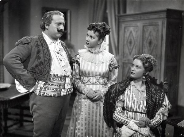 Scena del film "Elisir d'amore" - Regia Amleto Palermi, 1941- Jone Salinas, seduta a destra, osserva, insieme a un'attrice non identificata al centro, un attore non identificato a sinistra.