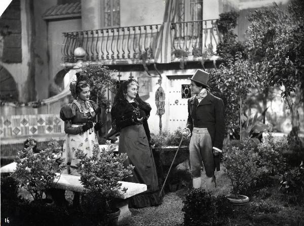 Scena del film "Elisir d'amore" - Regia Amleto Palermi, 1941- In un giardino, Pina Renzi, al centro, conversa con un attore non identificato. Intanto, sulla sinistra, un'attrice non identificata osserva la scena.