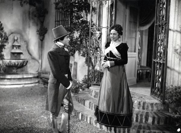 Scena del film "Elisir d'amore" - Regia Amleto Palermi, 1941- In un cortile, Margherita Carosio, a destra, guarda davanti a sè. Di fronte, un attore non identificato la guarda.