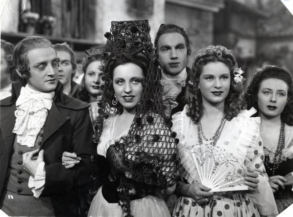 Scena del film "Elisir d'amore" - Regia Amleto Palermi, 1941- Al centro, Margherita Carosio, affiancata a destra da Jone Salinas, si appoggia al braccio di un attore non identificato. Alle loro spalle sono presenti altri attori non identificati.