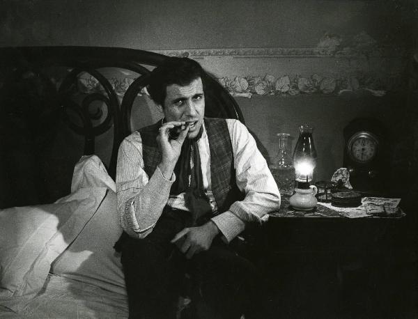 Scena del film "Er più storia d'amore e di coltello" - Regia Sergio Corbucci, 1971 - Adriano Celentano, seduto su un letto, guarda dritto davanti a sé mentre fuma un sigaro.