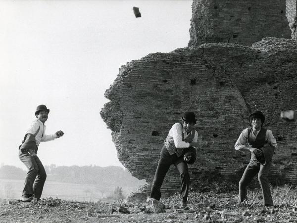 Scena del film "Er più storia d'amore e di coltello" - Regia Sergio Corbucci, 1971 - Ninetto Davoli, a destra, Adriano Celentano, al centro, e un attore non identificato lanciano delle pietre. Dietro, un muro e una fortezza diroccati.