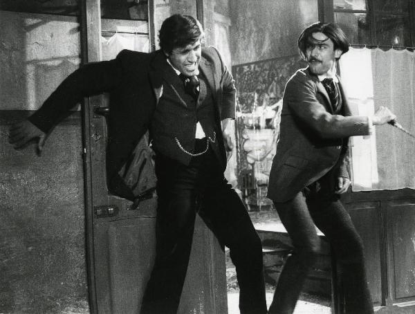 Scena del film "Er più storia d'amore e di coltello" - Regia Sergio Corbucci, 1971 - Adriano Celentano, a sinistra appoggiato a una porta aperta, si ripara dall'attacco di Gianni Macchia, a destra, che tiene in mano un coltello.