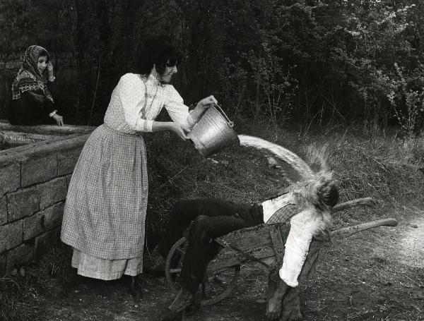 Scena del film "Er più storia d'amore e di coltello" - Regia Sergio Corbucci, 1971 - Claudia Mori getta un secchio d'acqua sul volto di Adriano Celentano. Intanto, una bambina seduta su un muretto in pietra, a sinistra, osserva la scena sorridendo.