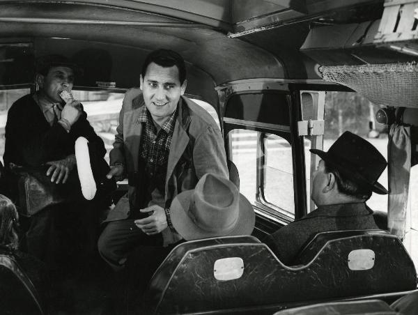 Scena del film "Era di venerdì 17" - Regia Mario Soldati, 1956 - Alberto Sordi, con un cappello da autista in mano, rivolge lo sguardo verso il fondo del pullmann. Intanto, un attore non identificato alle sue spalle lo osserva mangiando un panino.