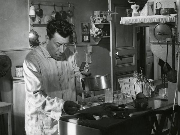 Scena del film "Era di venerdì 17" - Regia Mario Soldati, 1956 - In cucina: Fernandel che indossa un camice, è intento ad accendere con un fiammifero un fornello a gas mentre tiene nella mano sinistra un pentolino.