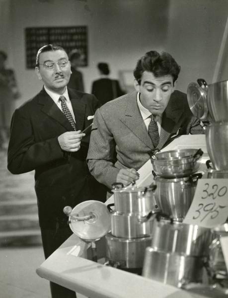 Scena del film "Era lui si si" - Regia Vittorio Metz, Marcello Marchesi, Marino Girolami, 1951 - Walter Chiari, a destra, osserva dietro una pile di pentole. Alle sue spalle lo guarda un attore non identificato che tiene una matita nella mano destra.