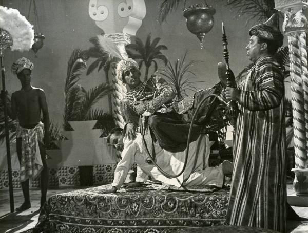 Scena del film "Era lui si si" - Regia Vittorio Metz, Marcello Marchesi, Marino Girolami, 1951 - Walter Chiari, vestito da sultano arabo, è steso sulla schiena di Carlo Campanini, e fuma un narghilè. Attorno, attori non identificati osservano.