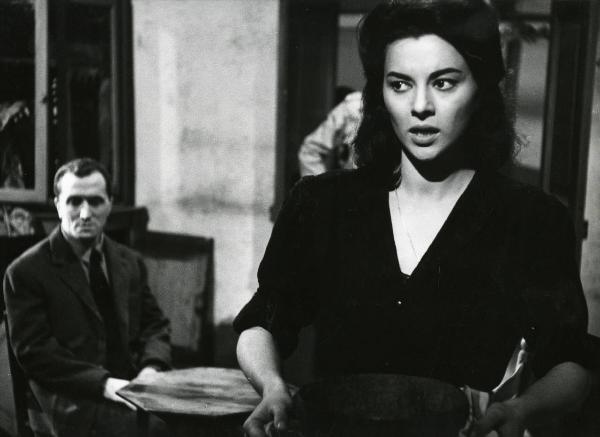 Scena del film "Era notte a Roma" - Regia Roberto Rossellini, 1960 - In primo piano, Giovanna Ralli volge lo sguardo verso sinistra. Alle sue spalle un attore non identificato, seduto a un tavolino, osserva verso il basso.