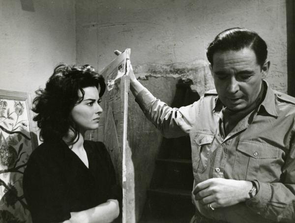 Scena del film "Era notte a Roma" - Regia Roberto Rossellini, 1960 - A sinistra, Giovanna Ralli a braccia conserte. A destra, Leon Genn appoggia la mano destra a una porta ricoperta di giornali.