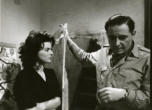 Scena del film "Era notte a Roma" - Regia Roberto Rossellini, 1960 - A sinistra, Giovanna Ralli a braccia conserte. A destra, Leon Genn appoggia la mano destra a una porta ricoperta di giornali.