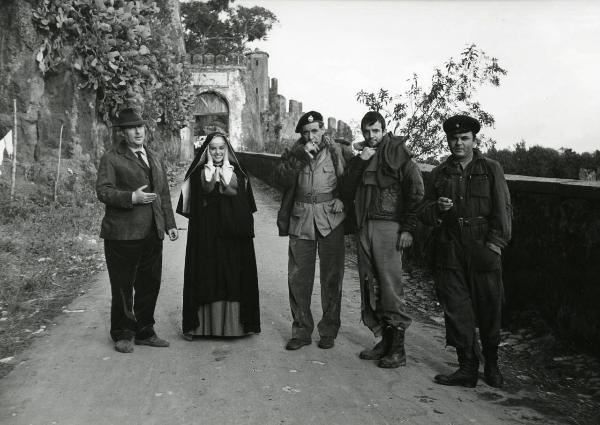 Scena del film "Era notte a Roma" - Regia Roberto Rossellini, 1960 - Figure intere di un attore non identificato, a sinistra, Giovanna Ralli, Leon Genn, al centro con una sigaretta in bocca, Peter Baldwin e Serghej Bondarciuk, a destra.