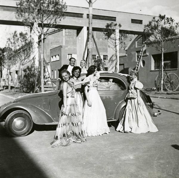 Scena del film "Eravamo 7 sorelle" - Regia Nunzio Malasomma, 1939 - Lotte Menas, a sinistra, con tre attrici non identificate, è in posa davanti a un'automobile in cui è seduto un attore non identificato. Dietro, Olivia Fried sorride a Nino Besozzi.