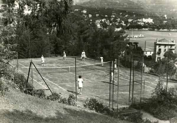 Scena del film "Eravamo 7 sorelle" - Regia Nunzio Malasomma, 1939 - Totale di un campo da tennis su cui sono intente a giocare sette attrici non identificate. In secondo piano sono visibili delle montagne, degli edifici e una distesa d'acqua.