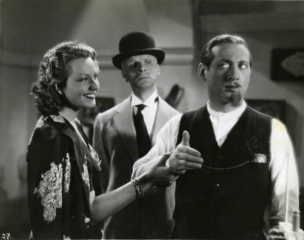 Scena del film "Eravamo 7 sorelle" - Regia Nunzio Malasomma, 1939 - Paola Barbara sorride mentre tiene il polso di Nino Besozzi, a destra con la barba e i baffi rasati per metà, che la guarda. Alle loro spalle un attore non identificato osserva .