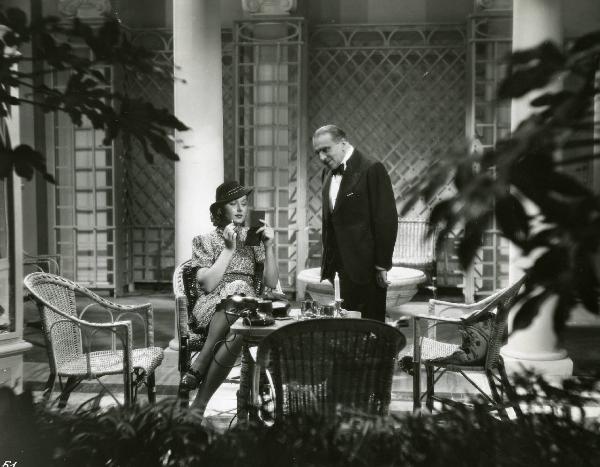 Scena del film "Eravamo 7 sorelle" - Regia Nunzio Malasomma, 1939 - Sergio Tofano, in piedi, osserva e sorride a Paola Barbara che, seduta a un tavolino, si trucca guardandosi in un piccolo specchio.