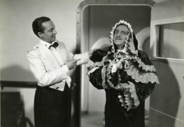 Scena del film "Eravamo sette vedove" - Regia Mario Mattoli, 1939 - Antonio Gandusio, a destra, è vestito con un abito con cappuccio decorato da merletti. Intanto, Nino Taranto, afferrandogli una spalla con la mano sinistra, lo indica e sorride.