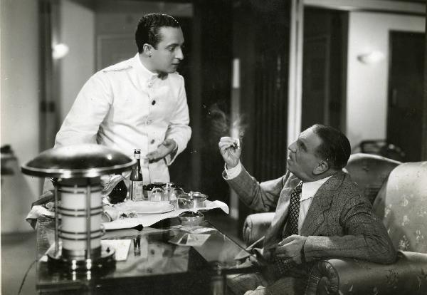 Scena del film "Eravamo sette vedove" - Regia Mario Mattoli, 1939 - A destra, Antonio Gandusio, seduto a una scrivania e con una sigaretta in mano, discute con Nino Taranto in piedi a sinistra, in vesti da cameriere.