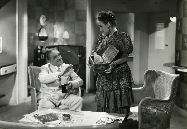 Scena del film "Eravamo sette vedove" - Regia Mario Mattoli, 1939 - Antonio Gandusio, seduto su una sedia, alza una mano mentre tiene con l'altra una cornice da tavolo. Intanto, un'attrice non identificata, lo guarda sorreggendo scatola.