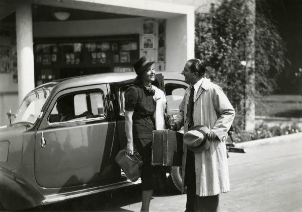 Scena del film "L'eredità in corsa" - Regia Oreste Biancoli, 1939 - Enrico Viarisio, con in mano un cappello e una valigia, guarda Clara Calamai, che, con una borsa in mano e una giacca appoggiata alla spalla sinistra, dà le spalle a un'automobile.