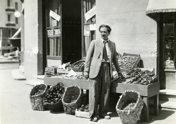 Scena del film "L'eredità in corsa" - Regia Oreste Biancoli, 1939 - Figura intera di Enrico Viarisio che tocca con la mano sinistra delle pere esposte davanti a un negozio di ortofrutta e rivolge lo sguardo verso sinistra.