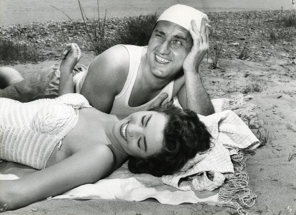 Scena del film "Un eroe dei nostri tempi" - Regia Mario Monicelli, 1955 - Alberto Sordi appoggia il capo alla mano sinistra, con cui tiene una sigaretta e rivolge lo sguardo sorridente verso sinistra, mentre Giovanna Ralli ride a occhi chiusi.