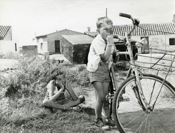 Scena del film "Eroe vagabondo" - Regia Walter Santesso, 1966 - Due piccoli attori non identificati guardano a destra. Uno è seduto a terra con le gambe incrociate e si tiene il viso tra le mani mentre l'altro sorregge una bicicletta.