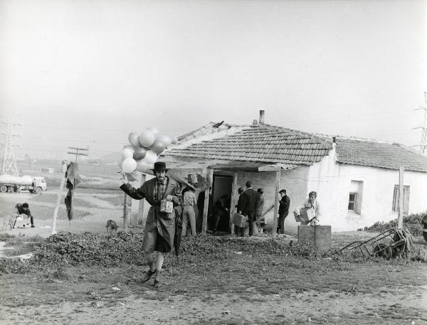 Scena del film "Eroe vagabondo" - Regia Walter Santesso, 1966 - Walter Santesso regge una scatola e osserva un attore non identificato che si dirige verso l'obbiettivo con dei palloncini e una scatola in mano. Dietro, attori non identificati.