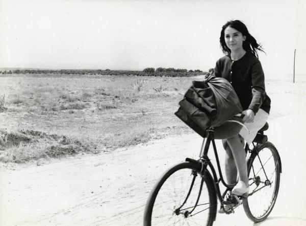 Scena del film "Eroe vagabondo" - Regia Walter Santesso, 1966 - Nuria Torray, in sella a una bici con cui trasporta un grande fagotto di stoffa, guarda verso l'obbiettivo e si dirige verso sinistra.