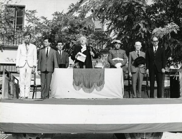 Scena del film "Eroe vagabondo" - Regia Walter Santesso, 1966 - Walter Santesso, a sinistra, rivolge lo sguardo verso il basso. Al centro, un attore non identificato parla al microfono e altri attori non identificati guardano davanti a loro.