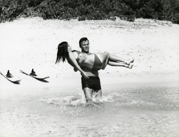 Scena del film "L'estate" - Regia Paolo Spinola, 1966 - Enrico Maria Salerno sostiene Mita Medici mentre entra in acqua. Entrambi sono in costume da bagno e alle loro spalle sulla spiaggia è presente un paio di sci.