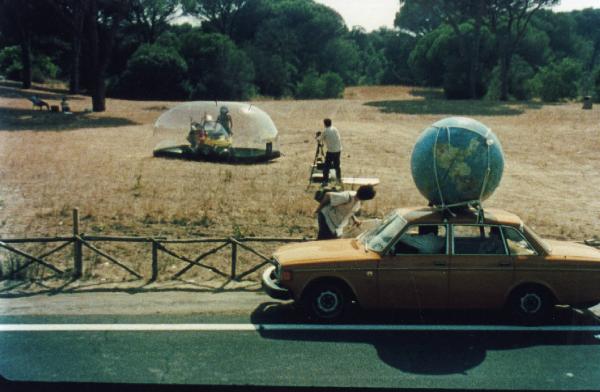 Sul set del film "Estate romana" - Regia Matteo Garrone, 2000 - Un operatore di spalle fotografa un gonfiabile con all'interno una vettura. In primo piano due attori non identificati sono dentro una macchina con un mappamondo legato al tetto.