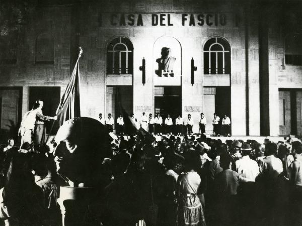 Scena del film "Estate violenta" - Regia Valerio Zurlini, 1959 - Attori non identificati osservano in direzione di un edificio recante la scritta CASA DEL FASCIO. All'ingresso è schierata una fila di attori non identificasti che guardano la folla.