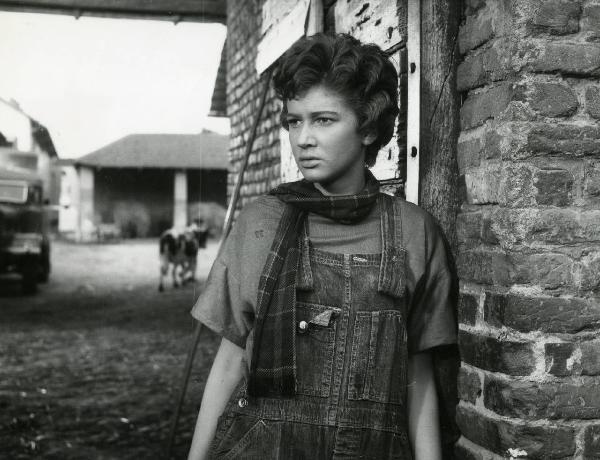 Scena del film "Esterina" - Regia Carlo Lizzani, 1959 - Mezza figura di Carla Gravina che indossa una salopette di jeans e una sciarpa al collo mentre rivolge lo sguardo verso sinistra.