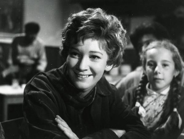 Scena del film "Esterina" - Regia Carlo Lizzani, 1959 - Primo piano di Carla Gravina che, a braccia conserte, sorride e rivolge lo sguardo verso sinistra. Alle sue spalle, attori e attrici non identificati.